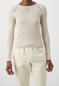 Ljusbeige ribbade långärmade topp med dekorativa utskärningar, ihopparad med krämfärgad byxor på en stående person mot vit bakgrund.