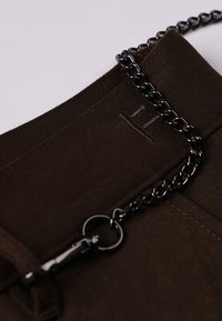 Tissu marron avec une finition texturée, comprenant une courte chaîne noire et un clip. Détail de couture discret visible près du haut.