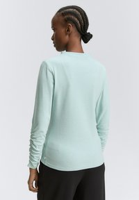 Kobieta z plecionymi włosami spiętymi w kok, ubrana w jasnoniebieską bluzkę z długim rękawem i marszczonymi rękawami oraz czarne spodnie, stoi tyłem do kamery.