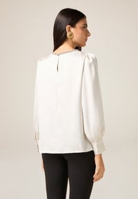 Weiße Bluse mit gepufften Ärmeln, geknöpften Manschetten, Schlüssellochdetail am Rücken und dekorativem Besatz am Halsausschnitt. Glatter, eleganter Stoff.