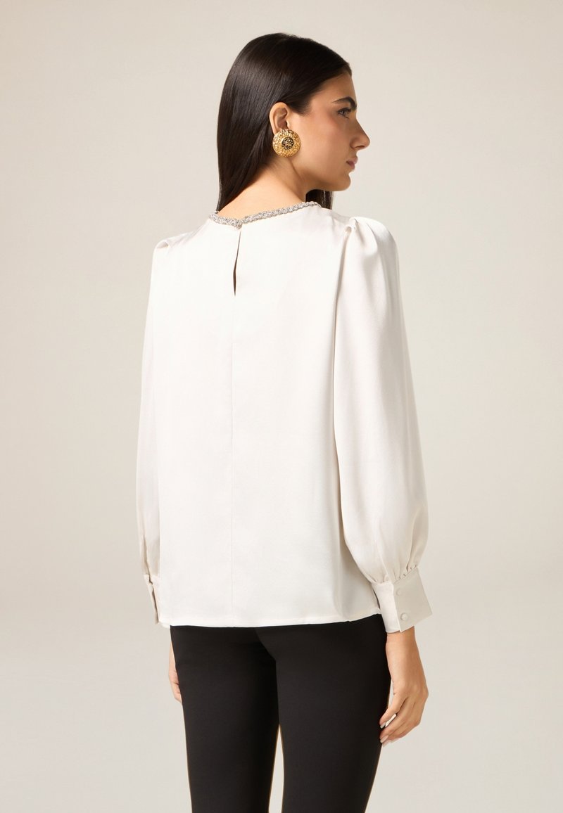 Weiße Bluse mit gepufften Ärmeln, geknöpften Manschetten, Schlüssellochdetail am Rücken und dekorativem Besatz am Halsausschnitt. Glatter, eleganter Stoff.