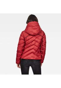 Veste matelassée rouge avec capuche, design à chevrons et finition brillante. La veste possède des poignets élastiques et une taille légèrement cintrée.