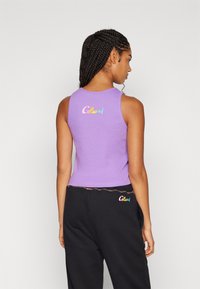 Lilla ermeløs crop topp med subtil ribbing; har "Colucci"-logo i gradientfarger på baksiden. Kombinert med svarte bukser.