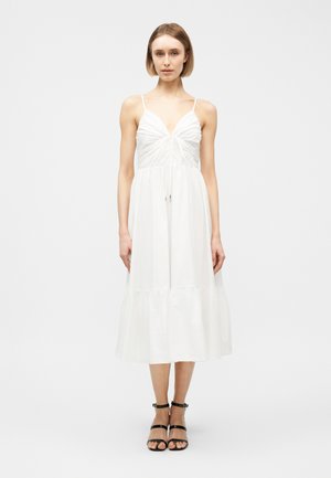 Vrouw die een witte mouwloze midi-jurk draagt met V-hals en geplooide buste, gecombineerd met zwarte sandalen met enkelbandje, staand tegen een effen achtergrond.