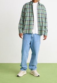 Grünes kariertes Hemd über einem weißen T-Shirt, kombiniert mit hellblauen weiten Jeans und beigen Sneakers. Merkmale sind eine lässige Passform und das Karomuster.