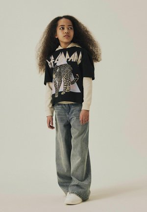 Camiseta gráfica negra con estampado de leopardo y el texto "Def Leppard" superpuesto a una camiseta de manga larga clara. Vaqueros sueltos de color azul claro y zapatillas blancas.
