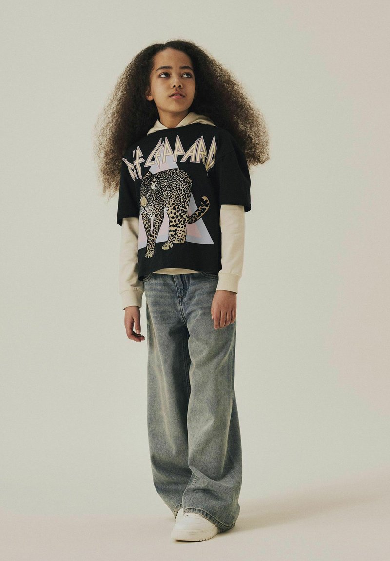 Svart grafisk t-shirt med leopardmönster och texten "Def Leppard" över en ljus långärmad tröja. Löst sittande ljusblå jeans och vita sneakers.