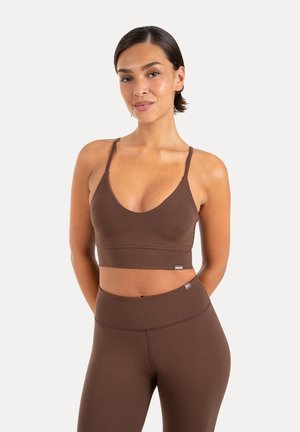 Femme debout, mains dans le dos, portant un brassière de sport marron et un legging taille haute assorti sur fond uni.