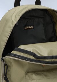 Interno di uno zaino in tela verde con fodera nera, dotato di una tasca con zip e un accento arancione. Cuciture rinforzate e spallacci regolabili.