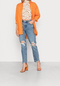 Camisa naranja de botones sobre una blusa estampada, jeans azules desgastados con detalles rasgados y sandalias de tacón bloque naranja.