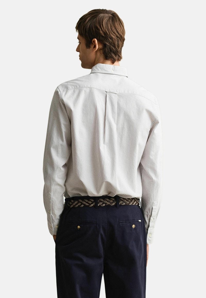 Mann, der ein hellgraues gestreiftes Langarmshirt trägt, in dunkelblaue Hosen mit geflochtenem braunem Gürtel gesteckt, von hinten gesehen.