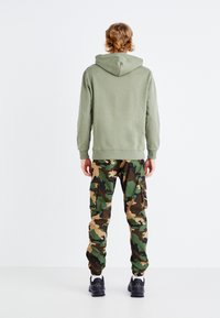 Felpa verde abbinata a pantaloni cargo con un motivo mimetico, dotati di diverse tasche e polsini elastici, indossati con sneaker nere.