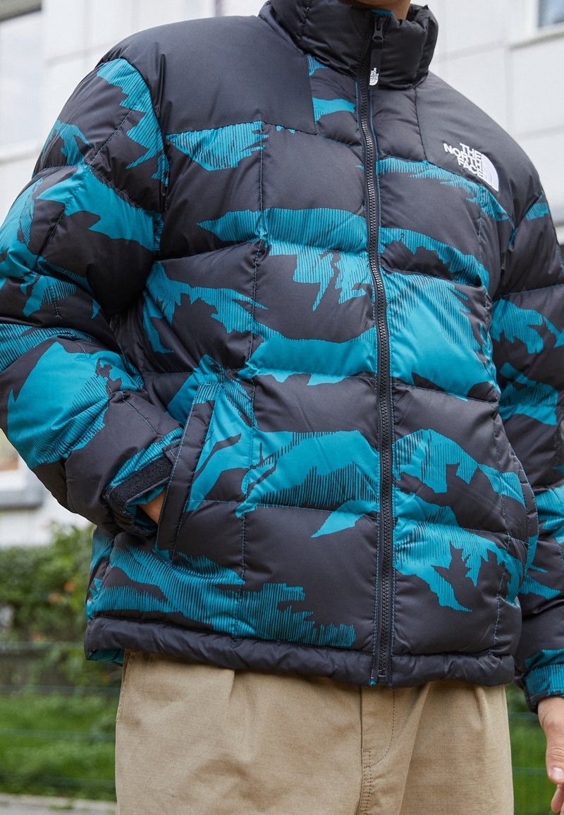 Giacca a vento imbottita con un motivo camouflage nero e teal, con chiusura a zip frontale, colletto alto e tessuto testurizzato, progettata per calore e comfort.
