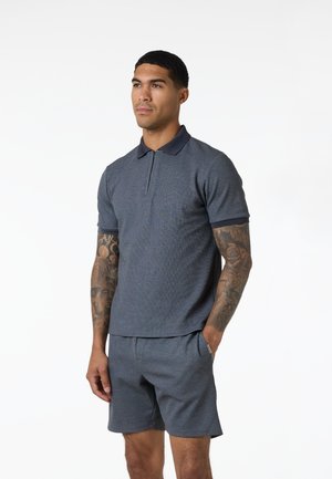 Mężczyzna z tatuażami na ramionach, ubrany w niebieską koszulkę polo z krótkim rękawem i pasujące krótkie spodenki, stoi z jedną ręką w kieszeni.