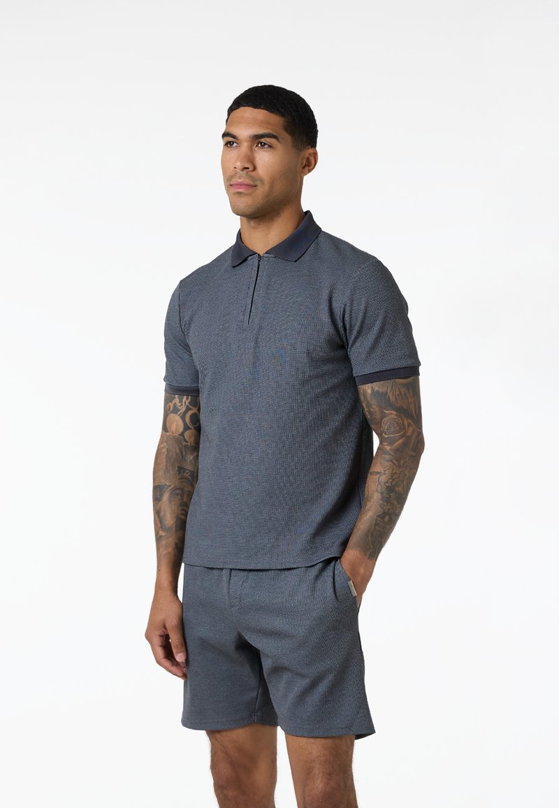 Homme aux bras tatoués portant un polo à manches courtes bleu et un short assorti, debout avec une main dans la poche.