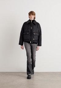 Casaco preto acolchoado com bolsos de pressão, apresentando um colarinho alto e um fecho de correr. Combinado com jeans cinzentos e calçado preto.