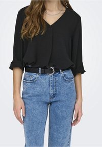 Blouse noire ample avec un col en V et des manches retroussées, associée à un jean en denim bleu clair taille haute et une ceinture noire avec une boucle en argent.