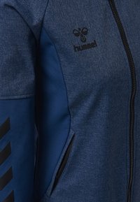 Ciemnoniebieska kurtka zapinana na zamek błyskawiczny z teksturowanej tkaniny, z czarnym logo na piersi i czarnymi ukośnymi paskami na rękawie.