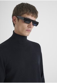 Crne okrugle sunčane naočale s debelim okvirom, nosi ih osoba u tamnom rolku puloveru. Glatka, sjajna površina, minimalno detalja.
