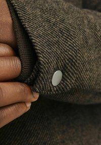 Ruskea houndstooth-kangas, jossa on teksturoitu pinta; sisältää hopeanapin; lähikuva kädestä, joka pitää hihaa.