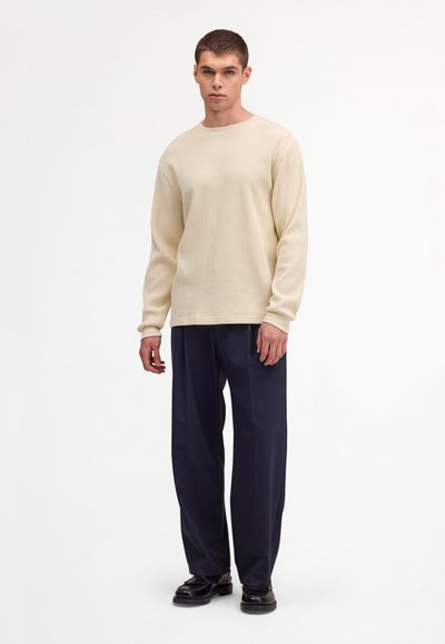 Jeune homme debout, portant un pull beige à manches longues texturé, un pantalon large bleu marine et des mocassins en cuir noirs, sur un fond blanc.
