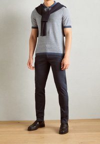 Polo in maglia con righe blu e bianche, abbinato a pantaloni blu scuro. Un maglione blu navale è adagiato sulle spalle. Sono indossate delle scarpe slip-on nere.