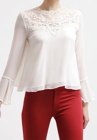 Blusa bianca con yoke in pizzo trasparente, ricami floreali, maniche lunghe a campana e orlo stratificato, abbinata a pantaloni rossi aderenti.