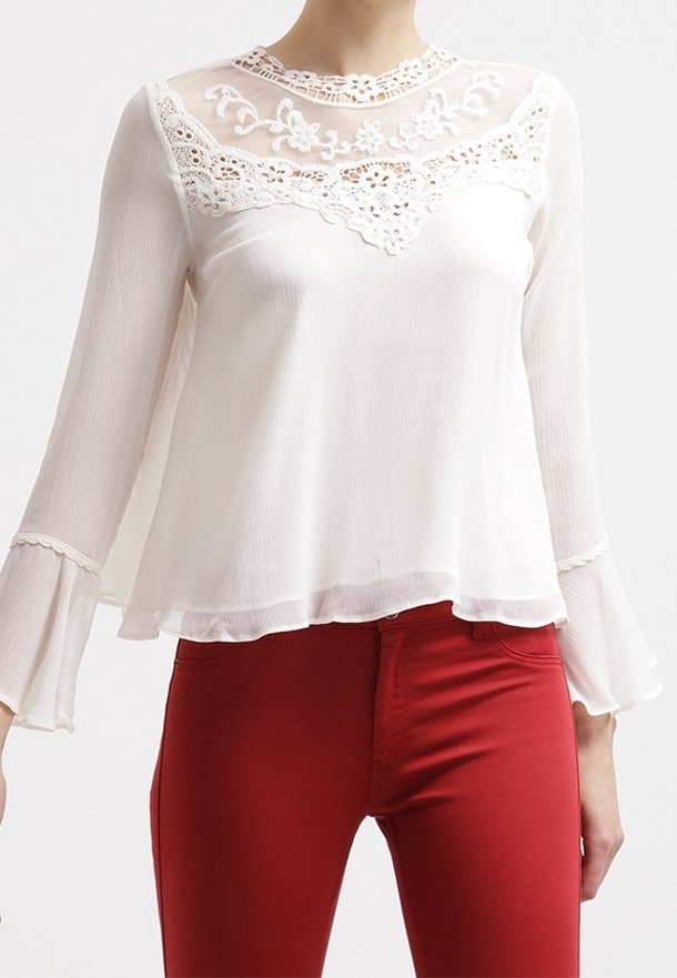 Blusa bianca con yoke in pizzo trasparente, ricami floreali, maniche lunghe a campana e orlo stratificato, abbinata a pantaloni rossi aderenti.
