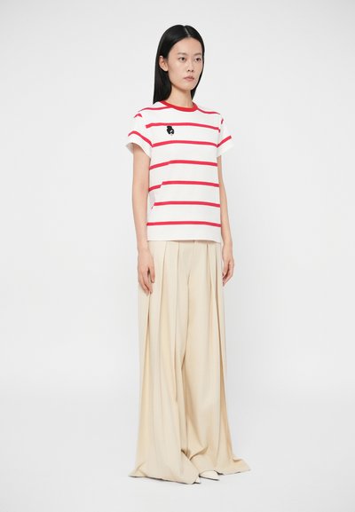 T-shirt branca com riscas vermelhas, mangas curtas e gráfico redondo preto no peito, acompanhada de calças largas bege claras.