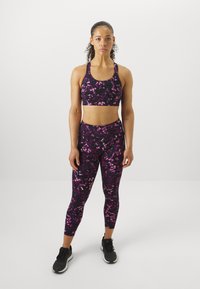 Sporto liemenėlė ir legginsai tamsios spalvos su violetiniais ir rožiniais gėlėmis. Prigludęs dizainas su lygiu paviršiumi. Juodi sportiniai batai.