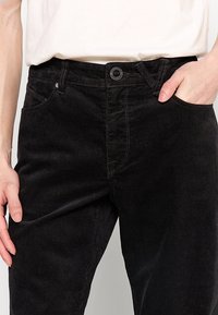 Gros plan d'une personne portant un pantalon en velours noir avec une main dans la poche et une chemise blanche rentrée.