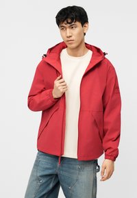 Jeune homme portant une veste à capuche rouge par-dessus un t-shirt blanc côtelé et un jean bleu, regardant légèrement sur le côté avec une expression neutre.