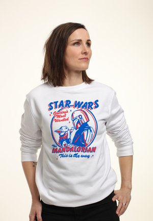 Kobieta w białej bluzie z kapturem z niebieskimi i czerwonymi grafikami z motywem Gwiezdnych Wojen - Mandalorianin i Grogu z tekstem "Galaxy's Most Wanted" oraz "This is the way."