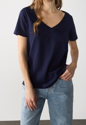 Vrouw met een marineblauw V-hals shirt met korte mouwen en lichtblauwe spijkerbroek, hand rustend in de voorzak.