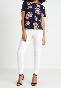 Navy floral short-sleeve top with pink flowers and green leaves paired with white fitted jeans and black strappy high-heeled sandals.