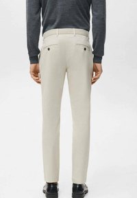 Mężczyzna ubrany w beżowe, dopasowane spodnie z tylnymi kieszeniami zapinanymi na guziki, ciemnoszarą koszulę z długim rękawem i czarne skórzane buty, stojący na białym tle.