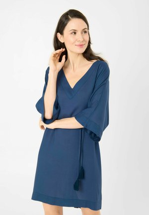 Frau mit langen dunklen Haaren trägt ein lockeres blaues Kleid mit weiten Ärmeln und einem fransigen Gürtel, blickt seitlich mit einem leichten Lächeln.