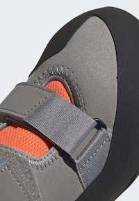 Chuteira de escalada cinza com um cabedal texturizado, forro laranja, tiras ajustáveis e sola em borracha. Apresenta costura reforçada e design ergonómico.