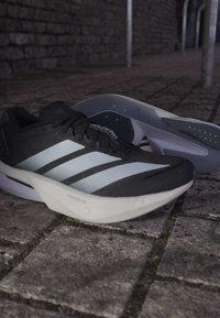 Sort-hvide Adidas løbesko med fremtrædende tre striber, placeret på en brostensbelagt gade.