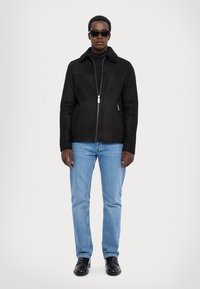 Schwarze Kunstwildlederjacke mit Schaffellkragen, Reißverschluss, kombiniert mit hellblauen Jeans und schwarzen Schuhen, vor einem neutralen Hintergrund.