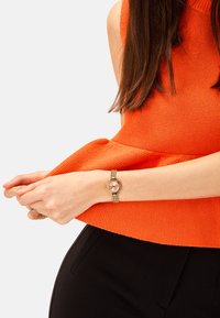 Gul metalur med en rund lyserød urskive, koblet band, båret på et håndled, parret med en orange strikket top og sorte bukser.