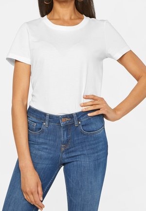 Woman wearing a plain white short-sleeve t-shirt tucked into high-waisted blue jeans, standing with one hand on her hip.