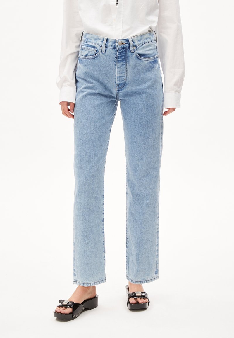 Femme portant un jean droit bleu clair, une chemise blanche à manches longues et des sandales plateformes noires avec des boucles décoratives.