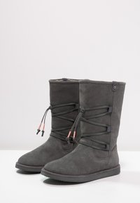Bottes en suede gris avec un design haut, présentant des lacets croisées et des accents de matériel rose en contraste sur le dessus. Semelle en caoutchouc plate.