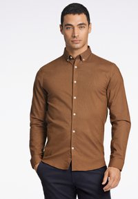 Camicia marrone a maniche lunghe con colletto button-down, tessuto testurizzato e bottoni bianchi, abbinata a pantaloni scuri.