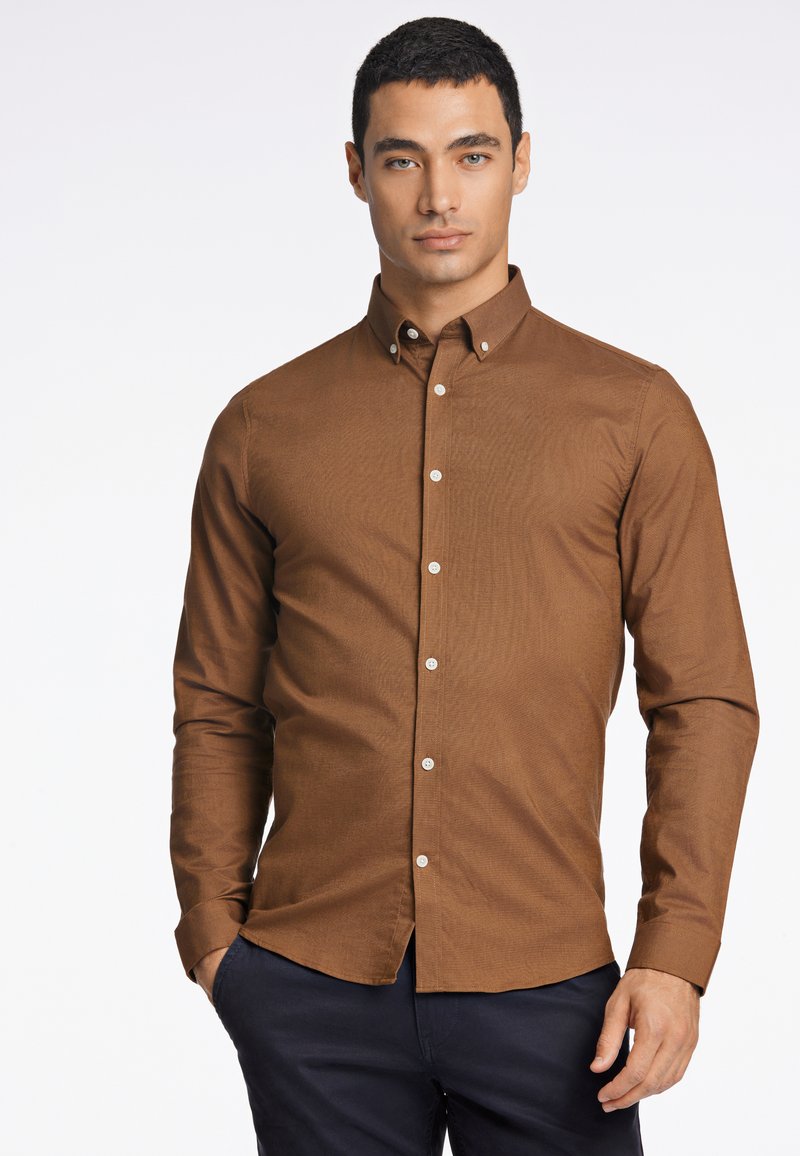 Camicia marrone a maniche lunghe con colletto button-down, tessuto testurizzato e bottoni bianchi, abbinata a pantaloni scuri.