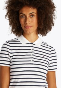 Kobieta o kręconych włosach w białej i granatowej koszulce polo w paski z małym logo na piersi, lekko się uśmiechająca na jednolitym tle.
