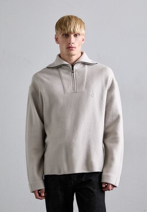 Lichtgrijze fleece pullover met een ritskraag, een detail op het voorpaneel en lange mouwen. De stof lijkt zacht met een gestructureerde afwerking.