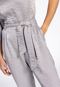 Grijze, gestructureerde broek met een paper-bag taille, voorzien van een strikceintuur, plooien en zijzakken. Soepele stof met een lichte glans.