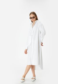 Weißes Hemdkleid mit langen Ärmeln, Seitentasche, asymmetrischem Saum, glatter Textur und lockerer Passform. Accessoires umfassen dunkle Sonnenbrillen.